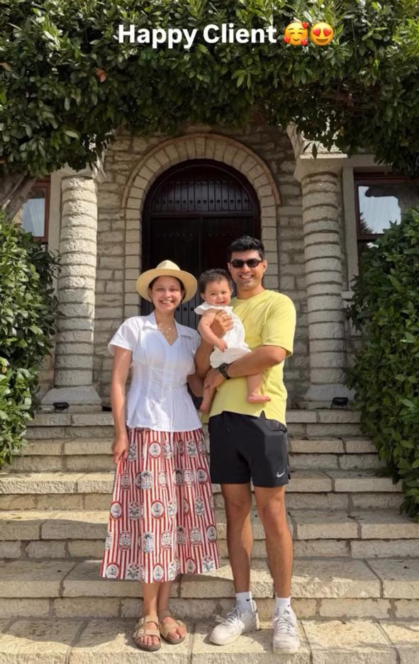 A family of three posing on stone steps in front of a stone building with green ivy. The woman wears a white top and a red-and-white patterned skirt, the man wears a yellow t-shirt and black shorts, and they are holding a baby.