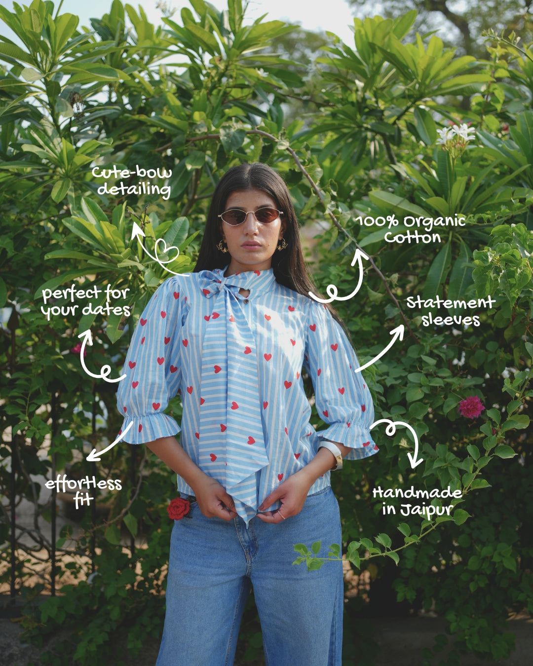 Woman wearing a blue striped shirt with heart patterns and jeans, surrounded by green foliage.