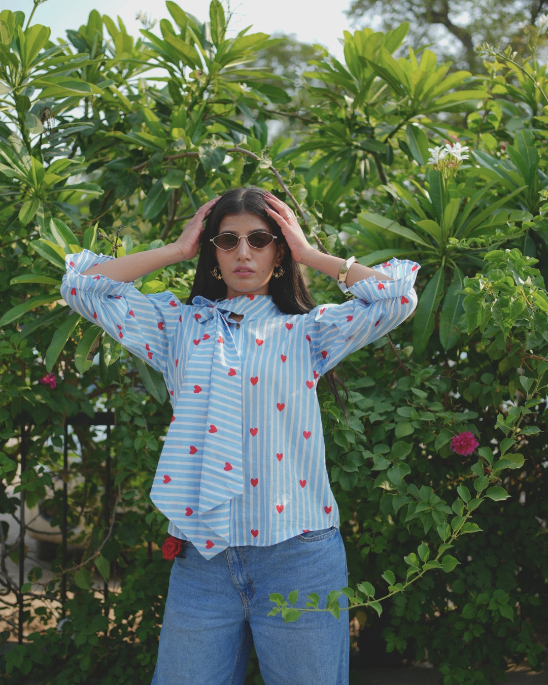 Person wearing a light blue shirt with red heart patterns and blue jeans, standing in front of green foliage.
