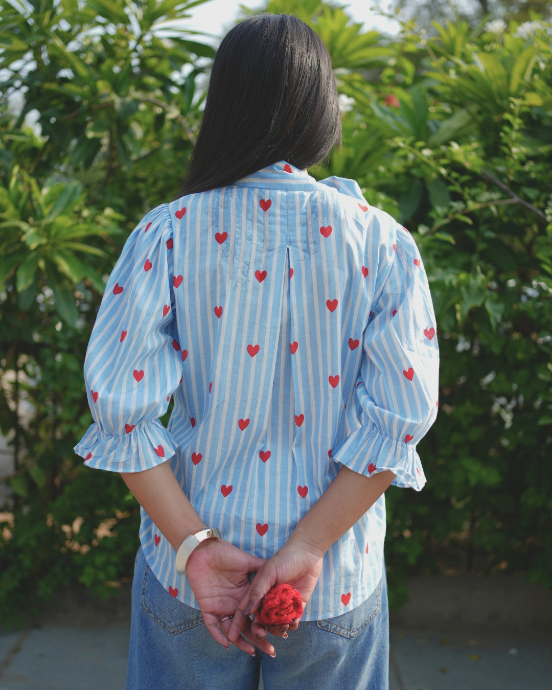 Person wearing a blue striped shirt with red heart patterns, standing outdoors with greenery in the background.