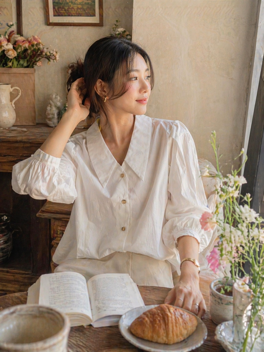 Woman in a white blouse sitting at a table with a book and bread, surrounded by flowers and a vase.