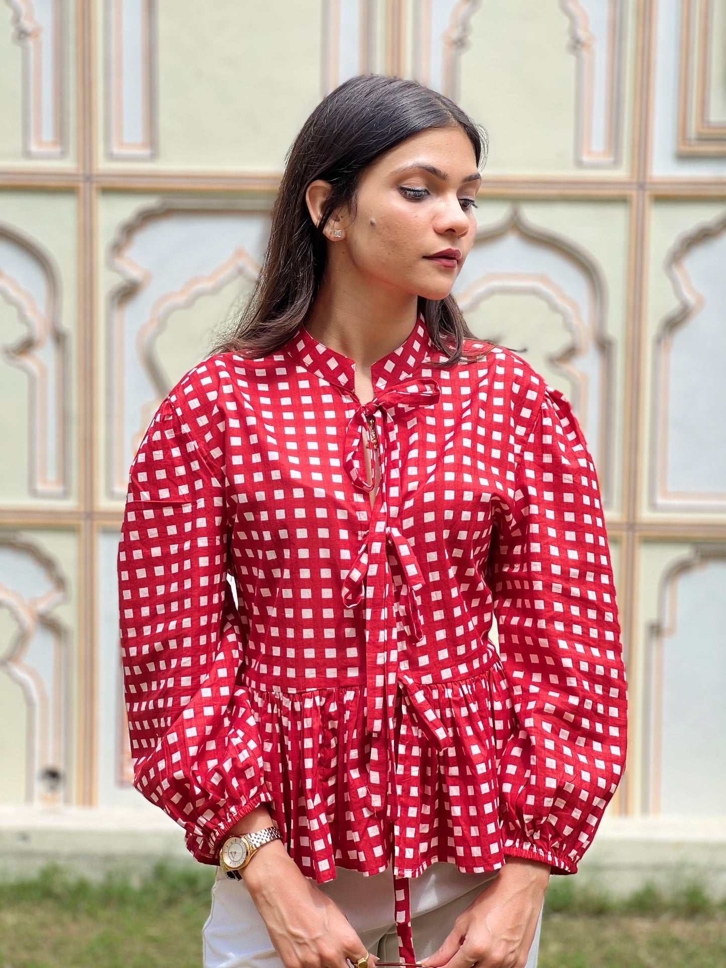 Woman wearing a red and white checkered blouse with a decorative pattern, standing in front of an ornate wall.