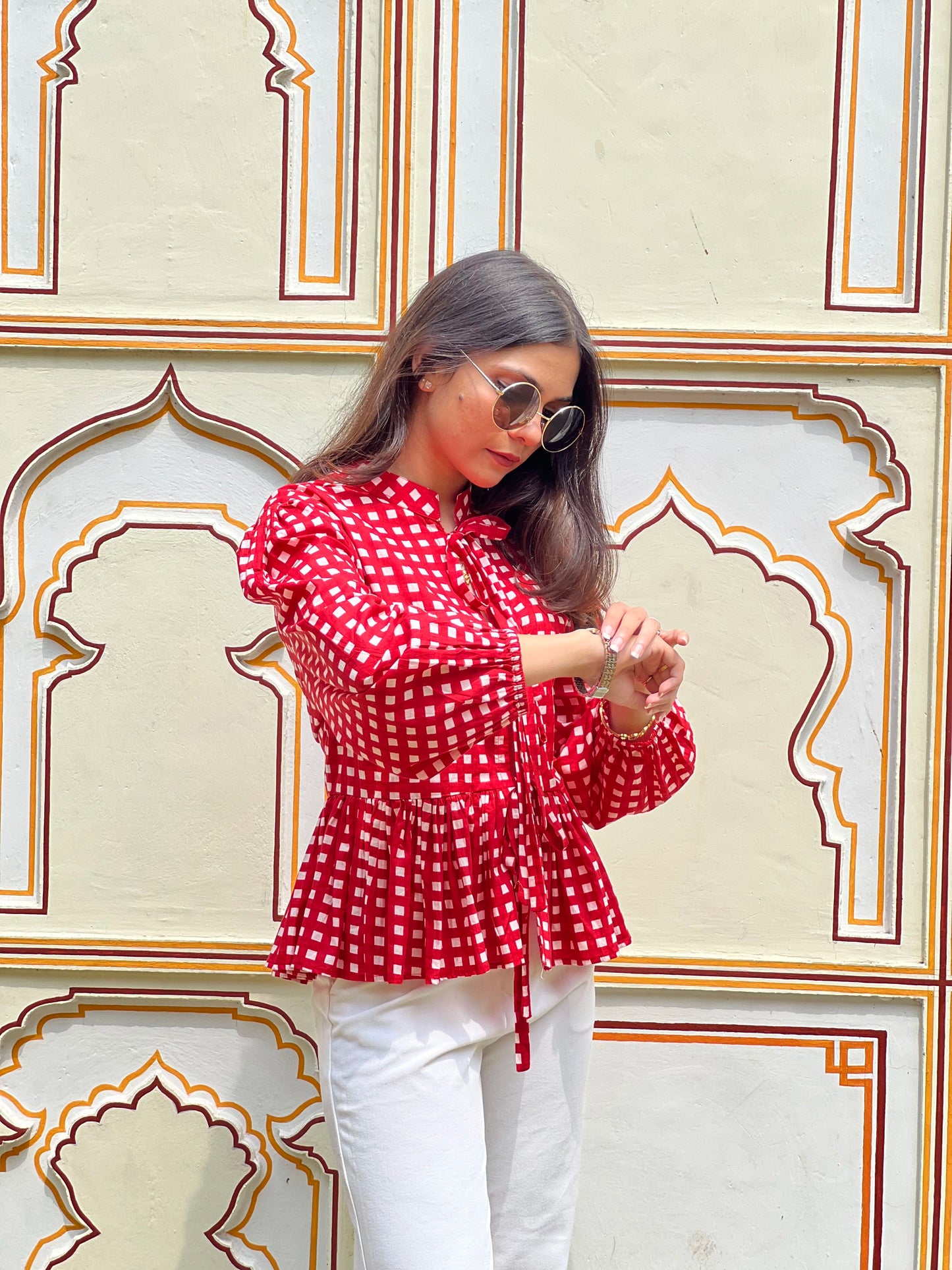 Woman wearing a red polka dot blouse and white pants against an ornate wall.