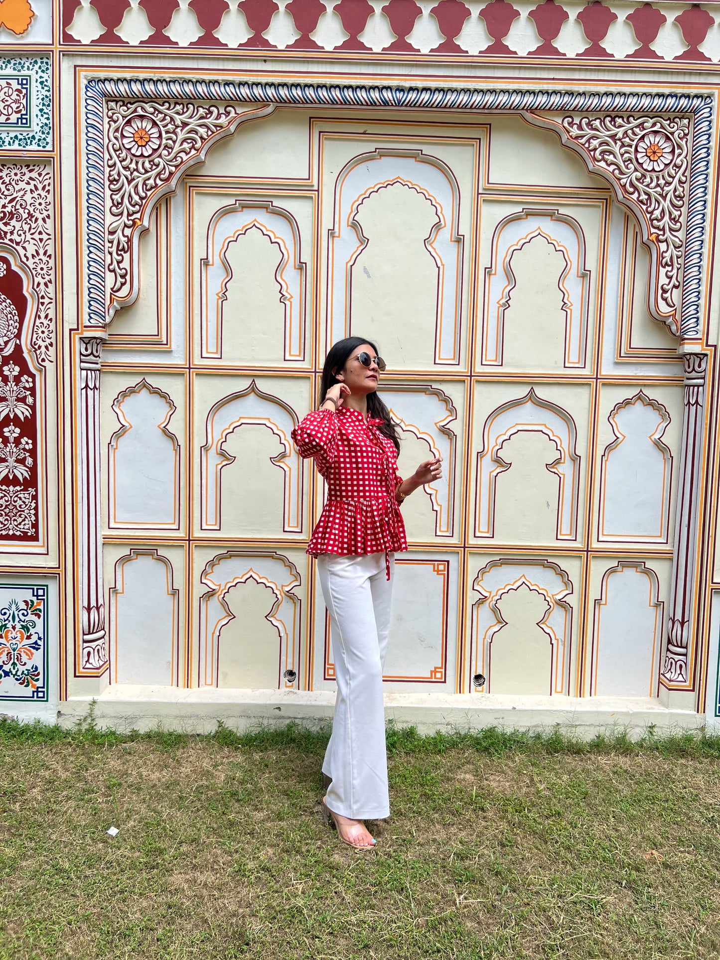 Woman in a red top and white pants standing in front of an ornate wall.