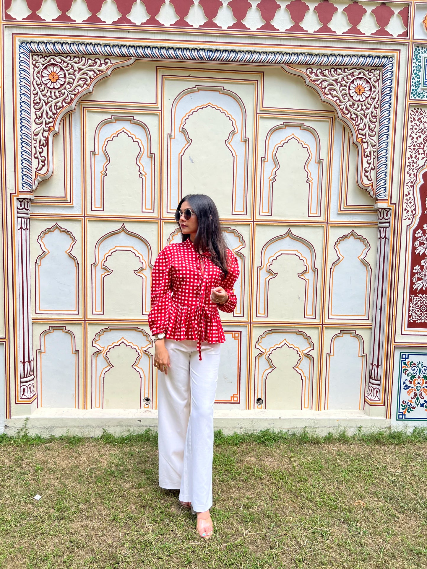Woman in a red checkered top and white pants standing in front of an ornate wall.