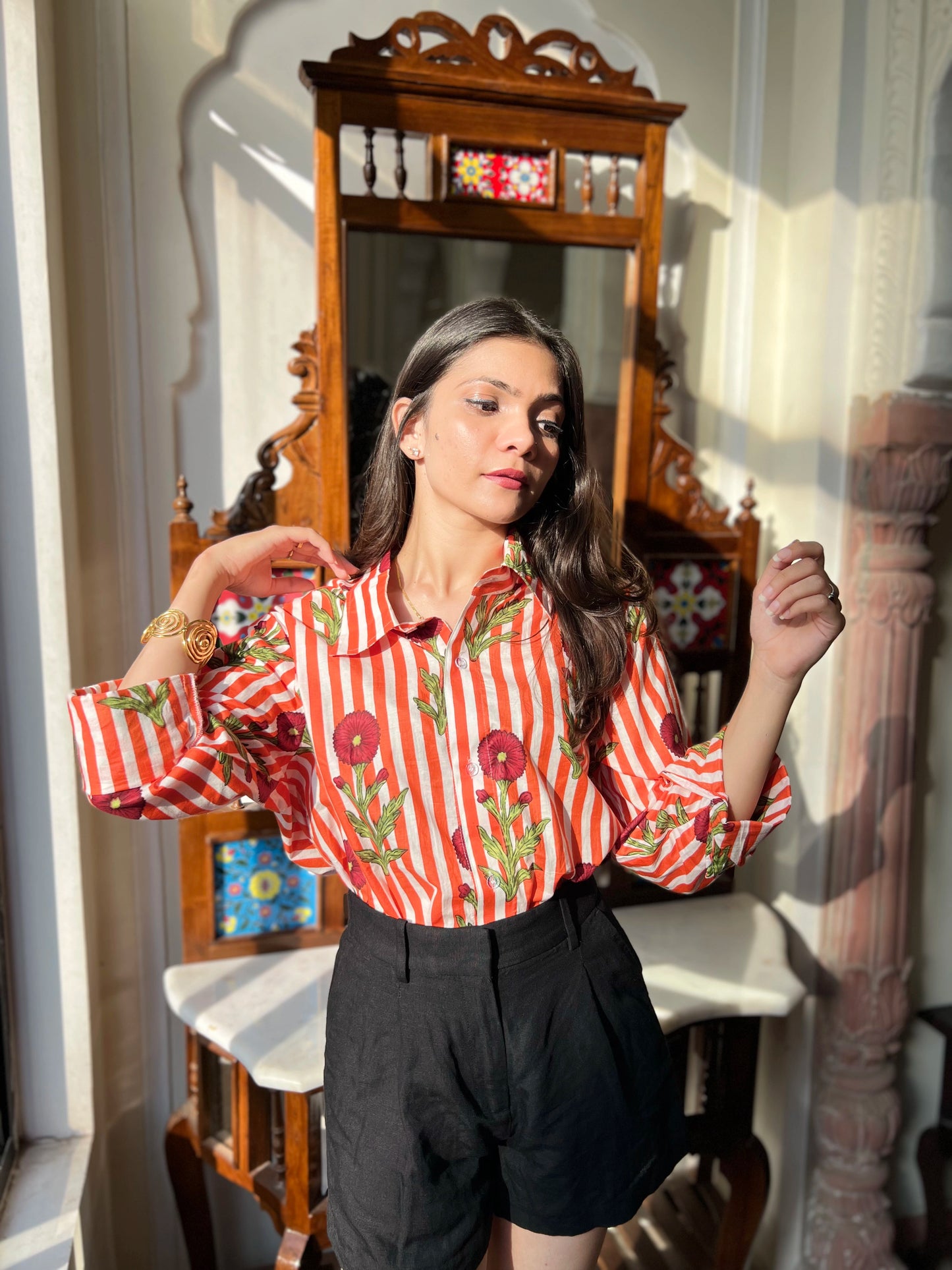 Woman wearing a red and white striped shirt with floral patterns in a room with a mirror and decorative items.