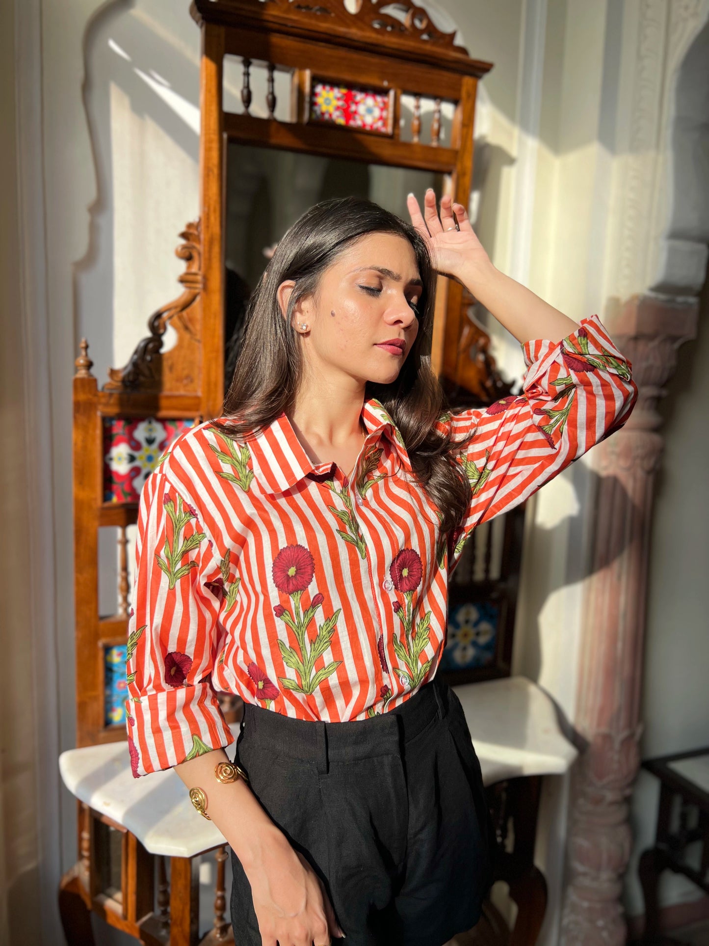 Woman wearing a red and white striped shirt with floral patterns in a room with a mirror and decor.