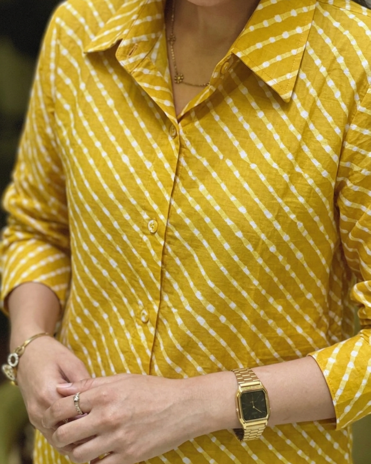 A close-up of a person wearing a yellow button-up shirt with a white diagonal dotted pattern, styled with gold jewelry including a watch, bracelet, and rings.